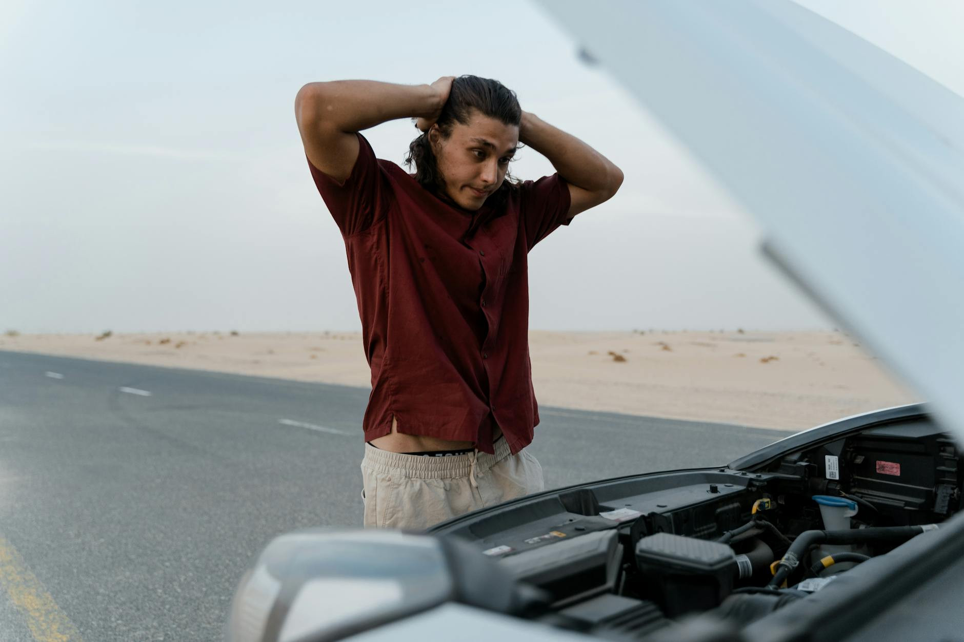 Un homme frustré examinant un moteur de voiture cassé sur une route déserte, montrant du stress et de la préoccupation.